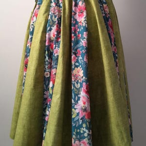 May include: A green skirt with a floral design. The skirt features a wide waistband and a full, flowing shape. The floral pattern showcases pink and red flowers on a dark teal background. The skirt is made of a textured fabric.