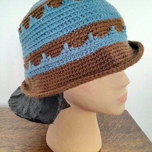 Puede incluir: Un gorro de punto marrón y azul con un ala ancha. El gorro tiene un patrón geométrico con rayas alternas de marrón y azul.