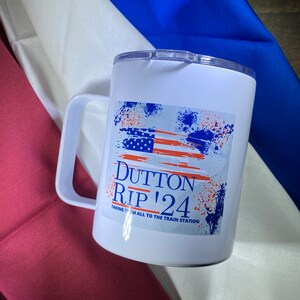 May include: White travel mug with a handle and a lid. The mug features a design with an American flag and the text "Dutton Rip '24". The design includes blue and orange paint splatters and the phrase "Taking them all to the train station."