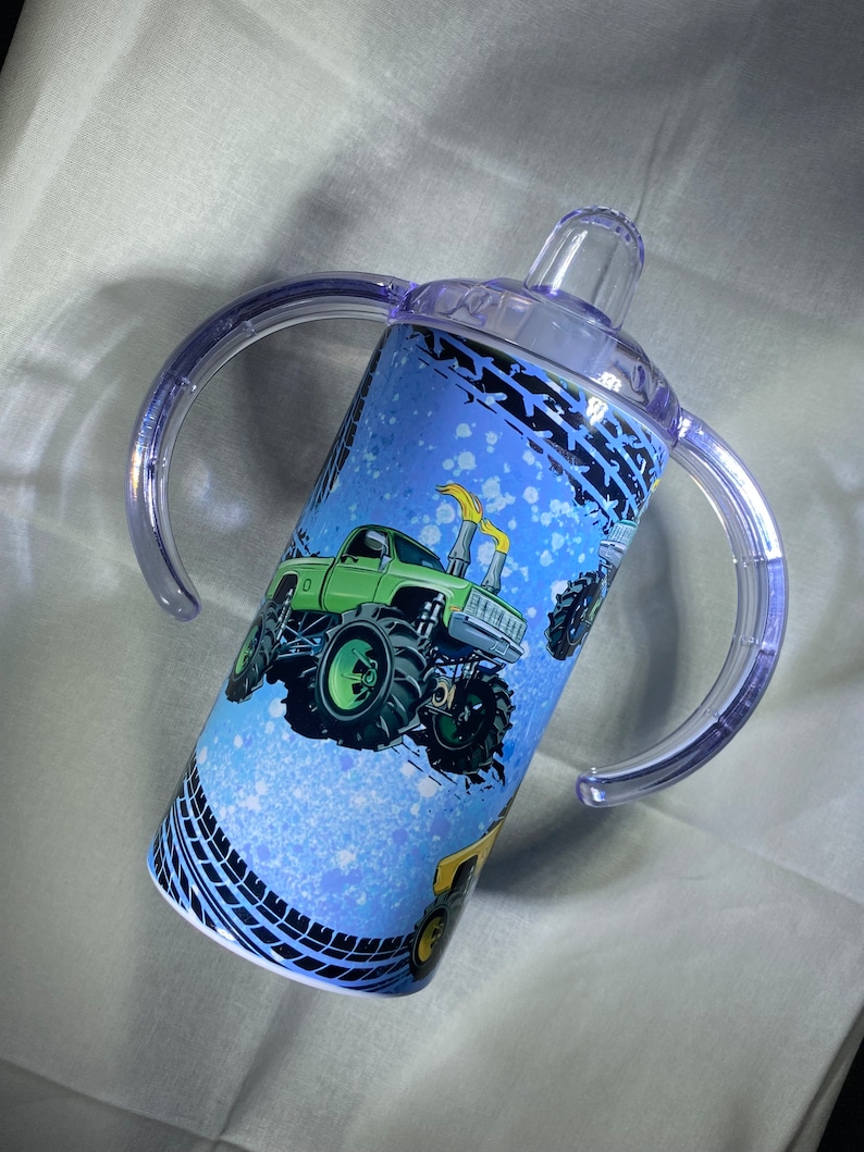 Puede incluir: Un vaso para beber con dos asas transparentes, con tem&aacute;tica de monster trucks azules y verdes. El vaso tiene una tapa transparente con un pico.