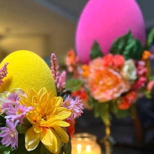 Może przedstawiać: Dwa świąteczne centralne elementy dekoracyjne z żółtymi i różowymi jajkami ozdobionymi kwiatami. Żółte jajko jest otoczone fioletowymi, różowymi i żółtymi kwiatami. Różowe jajko jest otoczone pomarańczowymi, różowymi i żółtymi kwiatami. Oba jajka znajdują się na ozdobnych podstawach.