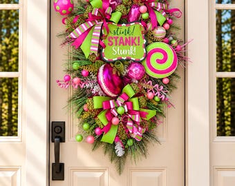 Op de afbeelding: Een druppelvormige krans met groene dennennaalden, felroze en limoengroene kerstballen, gestreepte linten en een centraal bord met de tekst "stink! STANK! Stunk!". De krans bevat een pluizige limoengroene elfenmuts en snoepversieringen.