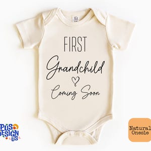 May include: A cream-colored onesie with the text "FIRST Grandchild Coming Soon" in black lettering. A small heart symbol is between the words "Grandchild" and "Coming".