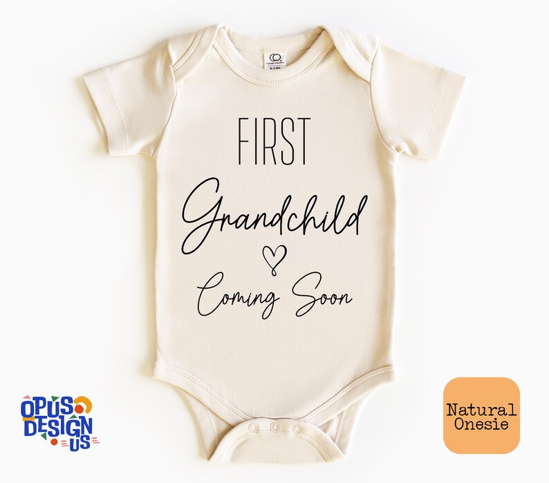 May include: A cream-colored onesie with the text "FIRST Grandchild Coming Soon" in black lettering. A small heart symbol is between the words "Grandchild" and "Coming".
