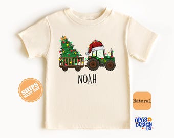 May include: A white t-shirt with a green tractor pulling a Christmas tree with lights. The tractor is wearing a Santa hat. The text "Noah" is printed below the tractor.