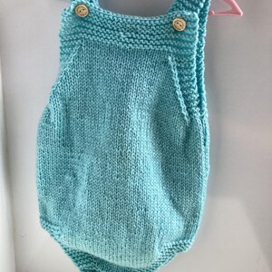 May include: A light blue knitted baby romper with wooden buttons on the straps. The romper is displayed on a pink hanger. The knitted fabric has a textured appearance, and the garment is designed with a rounded shape.