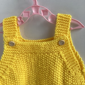 May include: A bright yellow knitted baby romper with wooden buttons on the straps. The garment is displayed on a pink plastic hanger against a plain white background. The knitted texture is clearly visible.