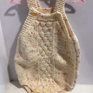 May include: A white knitted baby onesie with a speckled pattern and yellow buttons. The onesie has a cable knit design on the front and is made of a soft, comfortable material.