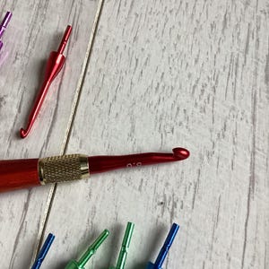 May include: A collection of colorful crochet hooks in various sizes. The hooks have metallic handles in red, green, blue, and purple. The hooks are arranged on a light wood-grain surface.