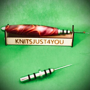 May include: A knitting needle with a red and brown marbled handle, resting in a wooden holder with the text "KNITSJUST4YOU". A second needle is also visible. The background is green.