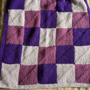 Könnte beinhalten: Eine gestrickte Babydecke mit einem karierten Muster in den Farben Lila, Rosa und Grau.