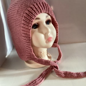 Op de afbeelding: Een roze gebreide muts met een puntige top en lange banden. De muts wordt getoond op een mannequinhoofd. De banden zijn onder de kin geknoopt. De muts is gemaakt van een zacht, getextureerd garen.