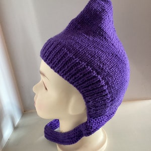 May include: A knitted purple hat with a long, pointed top and a neckband. The hat is displayed on a mannequin head, showcasing the texture and shape of the handmade item. The hat is a vibrant shade of purple.
