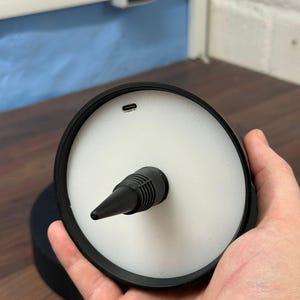 Op de afbeelding: Een zwarte oplaadstandaard met een wit rond oppervlak en een zwarte oplaadpoort. De standaard heeft een puntige punt aan de bovenkant.