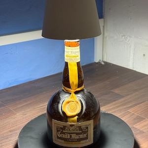 Op de afbeelding: Een hergebruikte Grand Marnier likeurfles met een geel lint en een waszegel, omgevormd tot een tafellamp met een witte lampenkap.