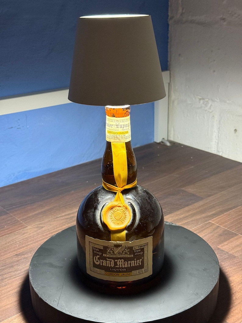 Op de afbeelding: Een hergebruikte Grand Marnier likeurfles met een geel lint en een waszegel, omgevormd tot een tafellamp met een witte lampenkap.