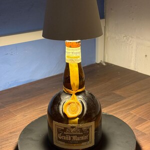 Op de afbeelding: Een hergebruikte Grand Marnier likeurfles met een witte lampenkap erboven. De fles is van bruin glas met een geel etiket waarop "Grand Marnier Liqueur Cordon Jaune" staat. De fles staat op een zwarte ronde voet.