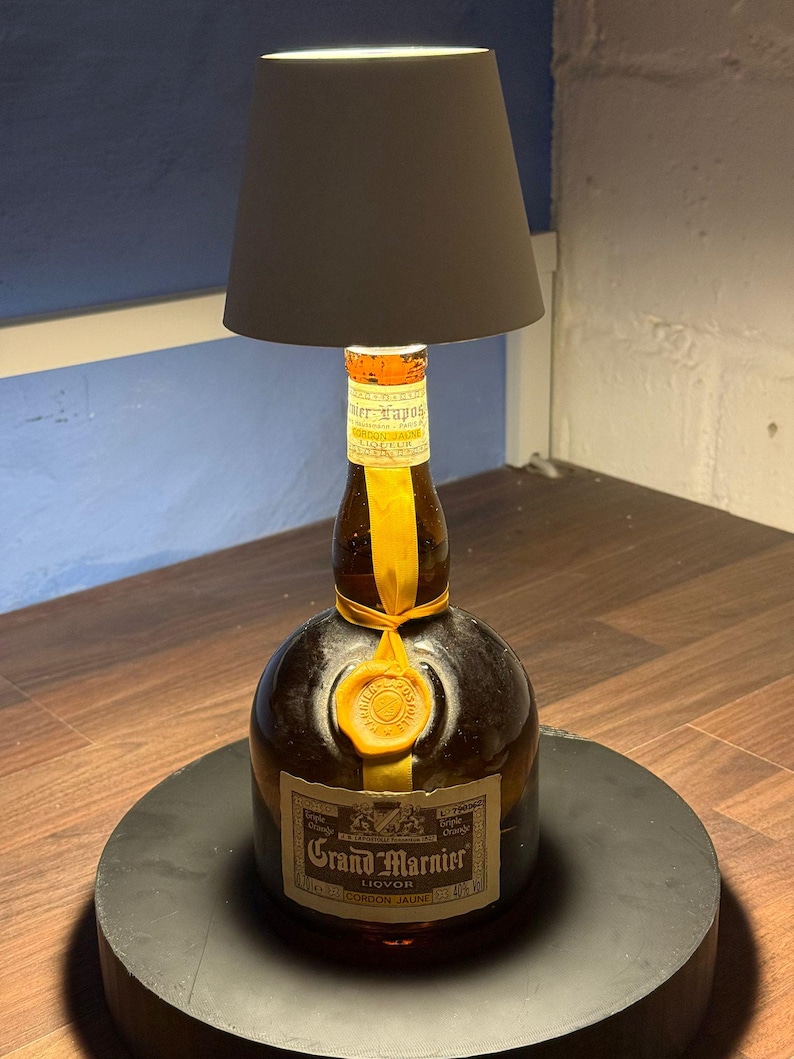 Op de afbeelding: Een hergebruikte Grand Marnier likeurfles met een witte lampenkap erboven. De fles is van bruin glas met een geel etiket waarop "Grand Marnier Liqueur Cordon Jaune" staat. De fles staat op een zwarte ronde voet.