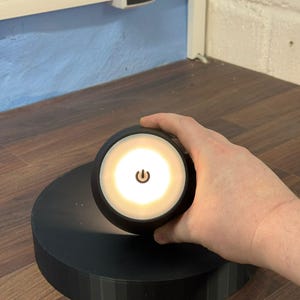 Op de afbeelding: Een zwarte, ronde, batterij-aangedreven LED-lamp met een aan/uit-knop aan de voorkant. Het licht is aan en verlicht de omgeving.