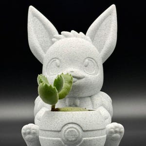 Può includere: Vaso da fiori in ceramica bianca a forma di animale dei cartoni animati con grandi orecchie e una piccola pianta che cresce al suo interno. Il vaso ha una base rotonda con una superficie testurizzata.