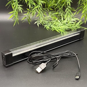 Könnte beinhalten: Ein schwarzer LED-Lichtbalken mit einer transparenten Kunststoffabdeckung, einem USB-Kabel und einem schwarzen rechteckigen Ende. Der Lichtbalken befindet sich auf einer schwarzen Oberfläche mit grünem Blattwerk im Hintergrund. Das USB-Kabel ist aufgerollt.