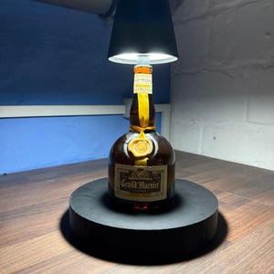 Op de afbeelding: Een zwarte lampenkap zit bovenop een fles Grand Marnier likeur. De fles staat op een zwarte ronde basis. De lampenkap is verlicht en geeft een warm licht.