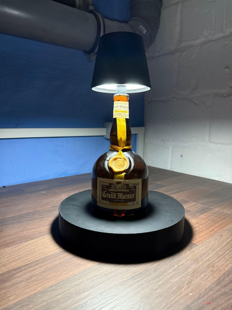 Op de afbeelding: Een zwarte lampenkap zit bovenop een fles Grand Marnier likeur. De fles staat op een zwarte ronde basis. De lampenkap is verlicht en geeft een warm licht.
