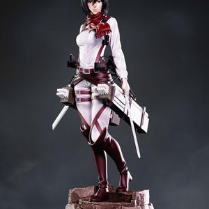 Pode incluir: Uma figura de estilo anime em escala 1:8 e 24 cm de altura, com cabelo preto curto, camisa branca de manga comprida, cachecol vermelho e equipamento de couro castanho com tiras. A figura segura duas lâminas prateadas sobre uma base rochosa.