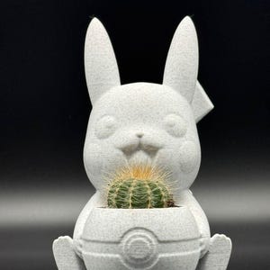 Peut inclure: Pot en céramique blanche en forme de lapin de dessin animé avec un petit cactus vert à l'intérieur. Le pot a une base ronde qui ressemble à une Poké Ball.