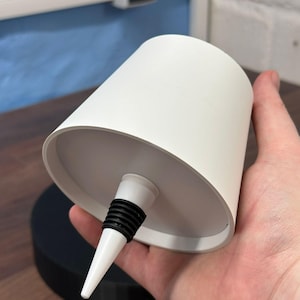 Op de afbeelding: Een witte keramische flessenstop met een zwarte rubberen ring en een puntige punt. De stop wordt in een hand gehouden.