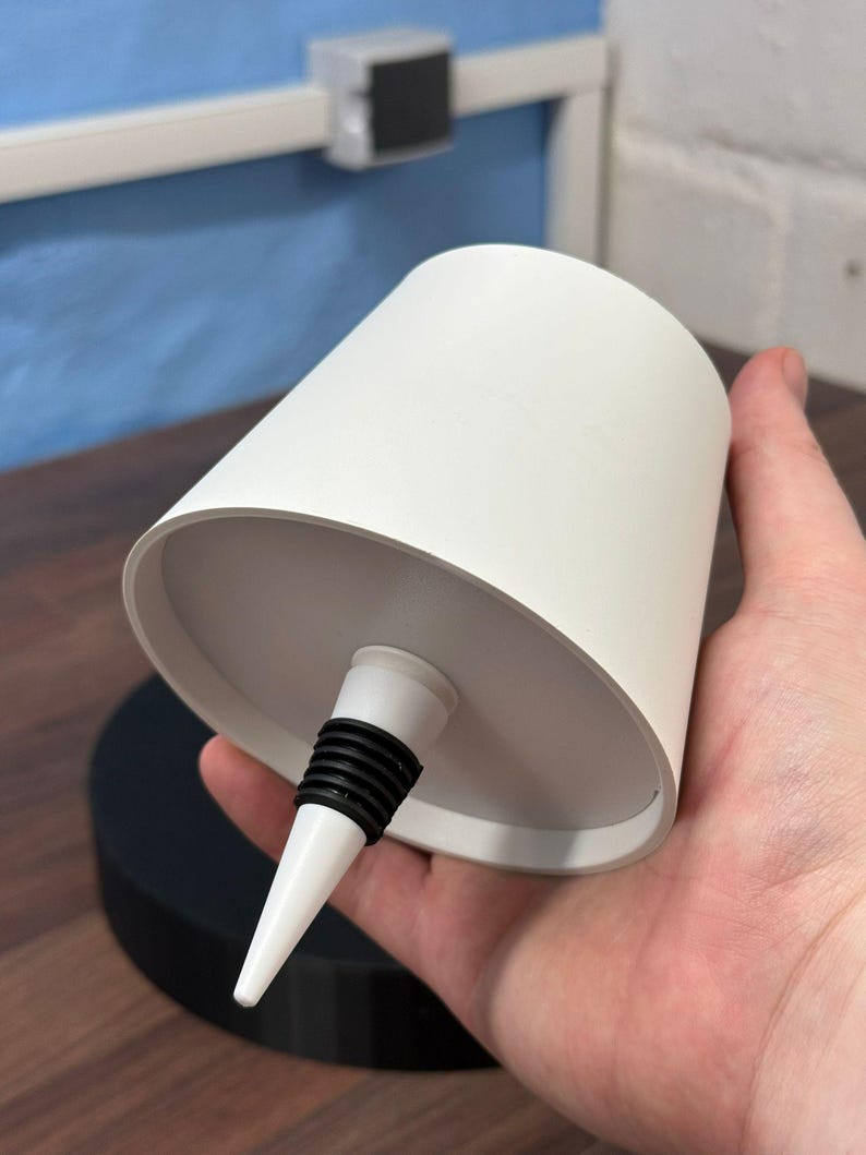Op de afbeelding: Een witte keramische flessenstop met een zwarte rubberen ring en een puntige punt. De stop wordt in een hand gehouden.