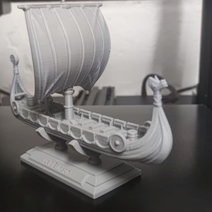 Könnte beinhalten: Ein graues 3D-gedrucktes Modell eines Wikingerschiffs mit einem Segel, einem Drachenkopf als Galionsfigur und einem Sockel mit dem Text "The Dragon".