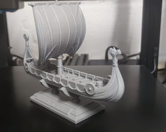 Op de afbeelding: Een grijs 3D-geprint model van een Vikingschip met een zeil, een drakenkop als boegbeeld en een basis met de tekst "The Dragon".