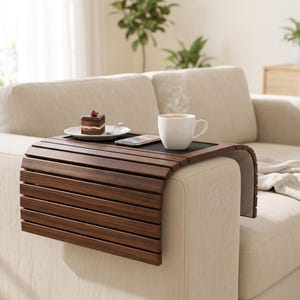 May include: A flexible dark brown wooden sofa arm tray with a flat black centre surface. The tray is draped over the arm of a beige couch and holds a white ceramic mug of steaming coffee, a smartphone, and a small slice of chocolate cake.