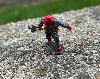 May include: A red and grey painted miniature figurine of a warrior holding an axe. The figurine is standing on a brown base.