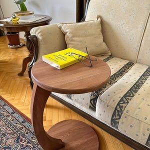 Peut inclure: Une table d'appoint en bois avec une jambe incurvée et un plateau rond. Un livre jaune avec le titre "The Savage God" est sur la table, ainsi qu'une paire de lunettes.