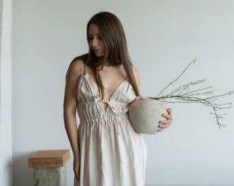 May include: A beige linen dress with spaghetti straps and a gathered waist. A textured, light gray vase holds a sprig of greenery. The dress has a keyhole detail at the bust. The background is a white wall.