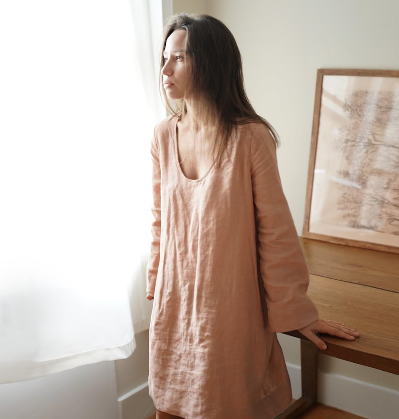Pode incluir: Vestido de linho cor de p&ecirc;ssego com mangas compridas e decote redondo. O vestido tem um corte folgado e vai at&eacute; meio da coxa. A modelo est&aacute; perto de uma janela e de uma mesa de madeira. O vestido &eacute; feito de tecido leve.