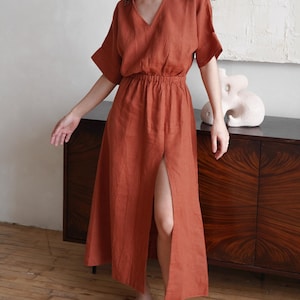 May include: Rust-colored linen dress with a V-neckline and short sleeves. The dress features an elastic waist and a high slit in the front. The dress is knee-length and made of a lightweight fabric.