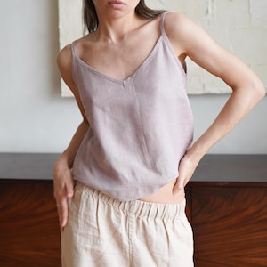 May include: A woman wearing a light pink linen tank top and beige linen pants. The tank top has a v-neck and thin straps.