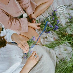 Pode incluir: Duas pessoas sentadas em uma manta arranjam uma grinalda floral. A cena inclui flores roxas, caules verdes, barbante azul e tesouras brancas. Uma pessoa usa um top rosa, a outra um top branco.