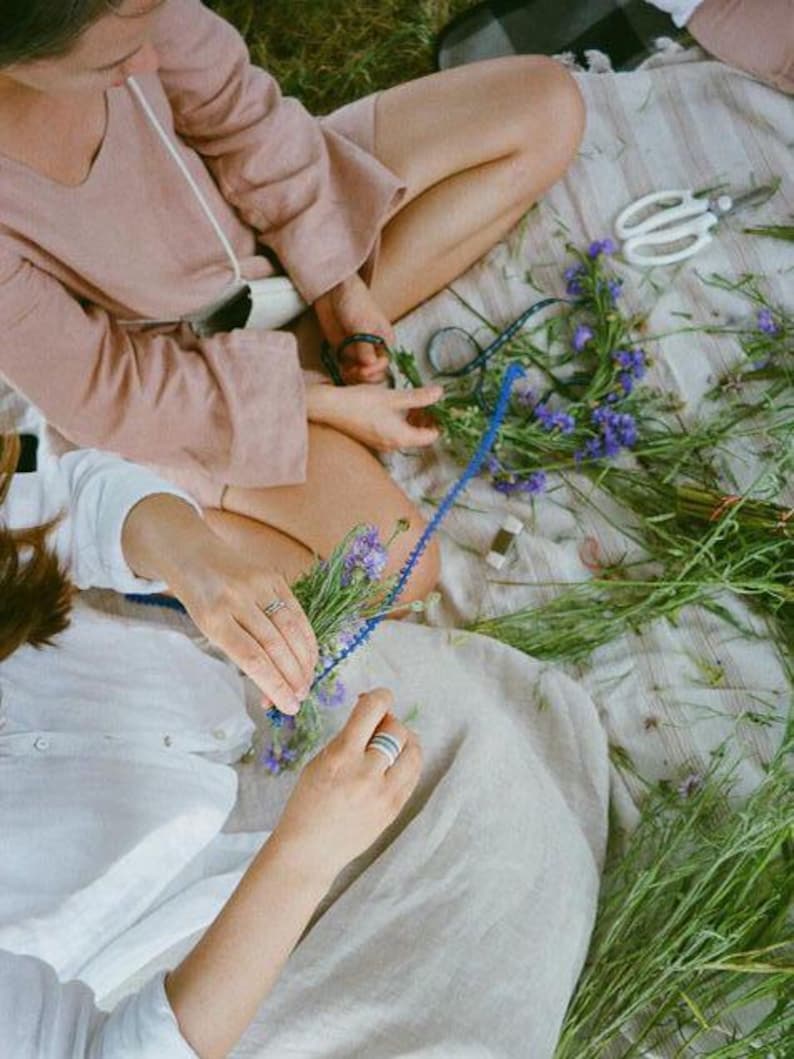 Pode incluir: Duas pessoas sentadas em uma manta arranjam uma grinalda floral. A cena inclui flores roxas, caules verdes, barbante azul e tesouras brancas. Uma pessoa usa um top rosa, a outra um top branco.