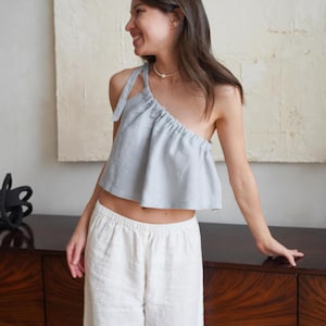 May include: A light blue one-shoulder top and white linen pants. The top has a gathered neckline and a single shoulder strap. The pants are wide-legged and have an elastic waistband. The outfit is made of a lightweight fabric.
