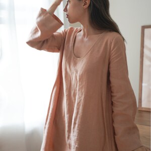 Pode incluir: Um vestido de linho cor de p&ecirc;ssego com mangas compridas e decote redondo. O vestido tem uma silhueta solta e fluida e mangas largas em sino. A modelo est&aacute; perto de uma janela.