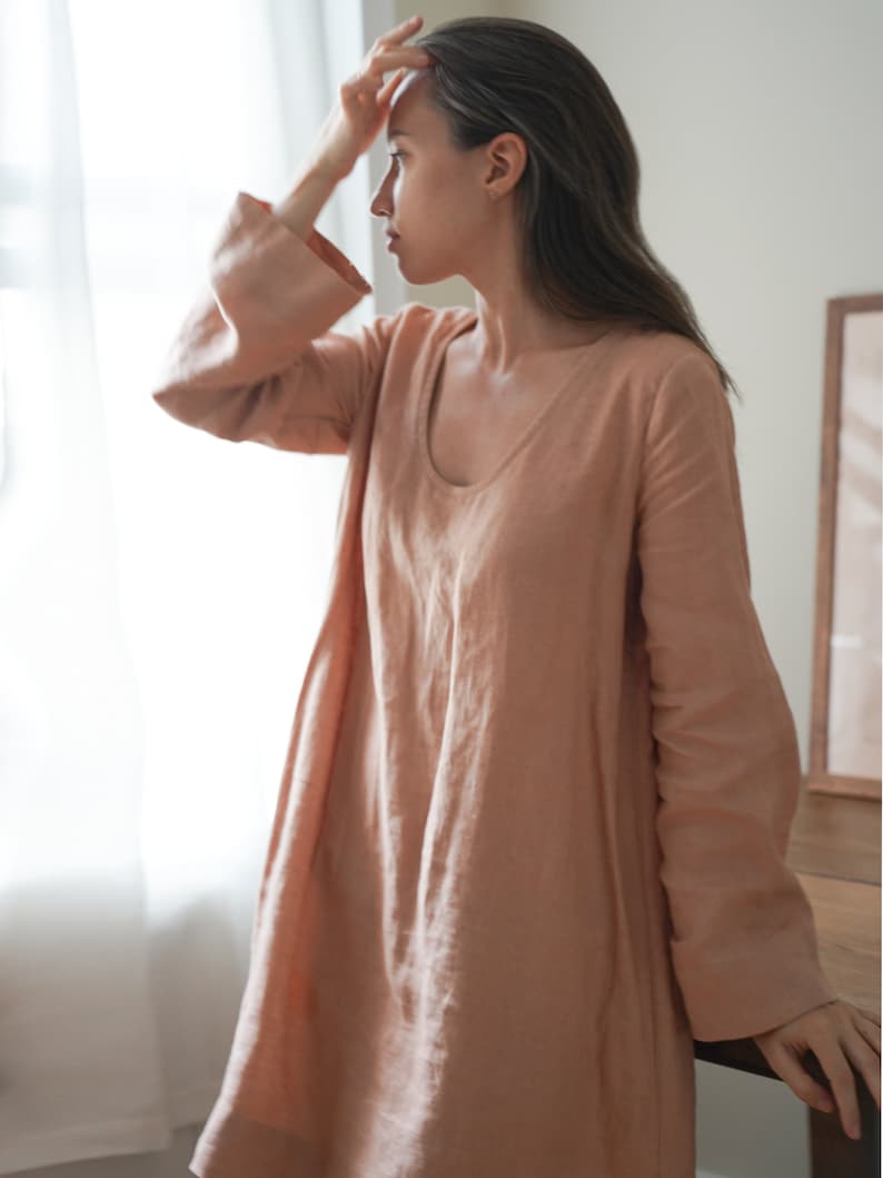 Pode incluir: Um vestido de linho cor de p&ecirc;ssego com mangas compridas e decote redondo. O vestido tem uma silhueta solta e fluida e mangas largas em sino. A modelo est&aacute; perto de uma janela.
