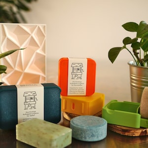 Peut inclure: Un ensemble de quatre contenants à savon réutilisables et colorés avec un motif de puits sur l'étiquette. Les contenants sont fabriqués à partir de plastique recyclé et sont conçus pour contenir des barres de shampoing solides, des barres de revitalisant et des barres de savon pour le corps. Les contenants sont présentés sur une surface en bois avec deux barres de savon.