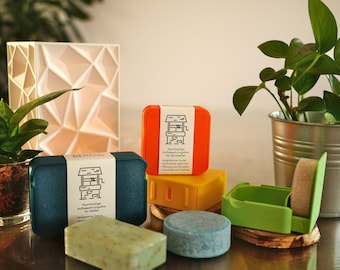 May include: A set of four colorful, reusable soap containers with a well design on the label. The containers are made of plastic and are designed to hold solid shampoo bars. The containers are sitting on a wooden surface with two solid shampoo bars.