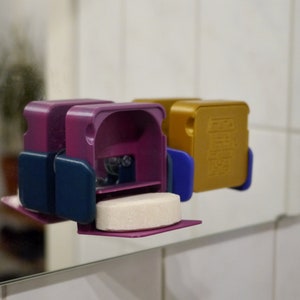 May include: Two 3D printed soap dispensers, one purple and one gold, with a white soap tablet in the purple dispenser. The dispensers have a blue accent on the side.