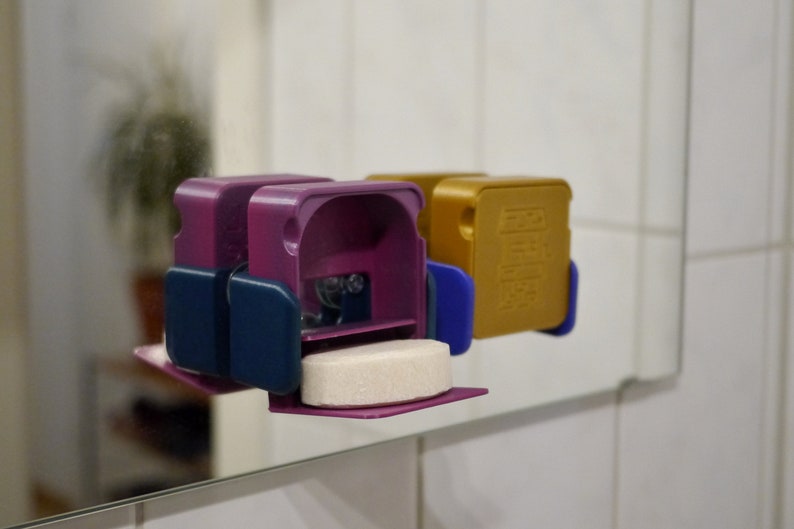 May include: Two 3D printed soap dispensers, one purple and one gold, with a white soap tablet in the purple dispenser. The dispensers have a blue accent on the side.