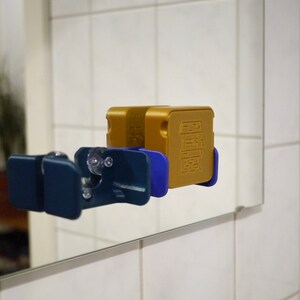 May include: Two gold-colored boxes with a design of a building on the front are attached to a blue and white wall mount. The wall mount is attached to a mirror.